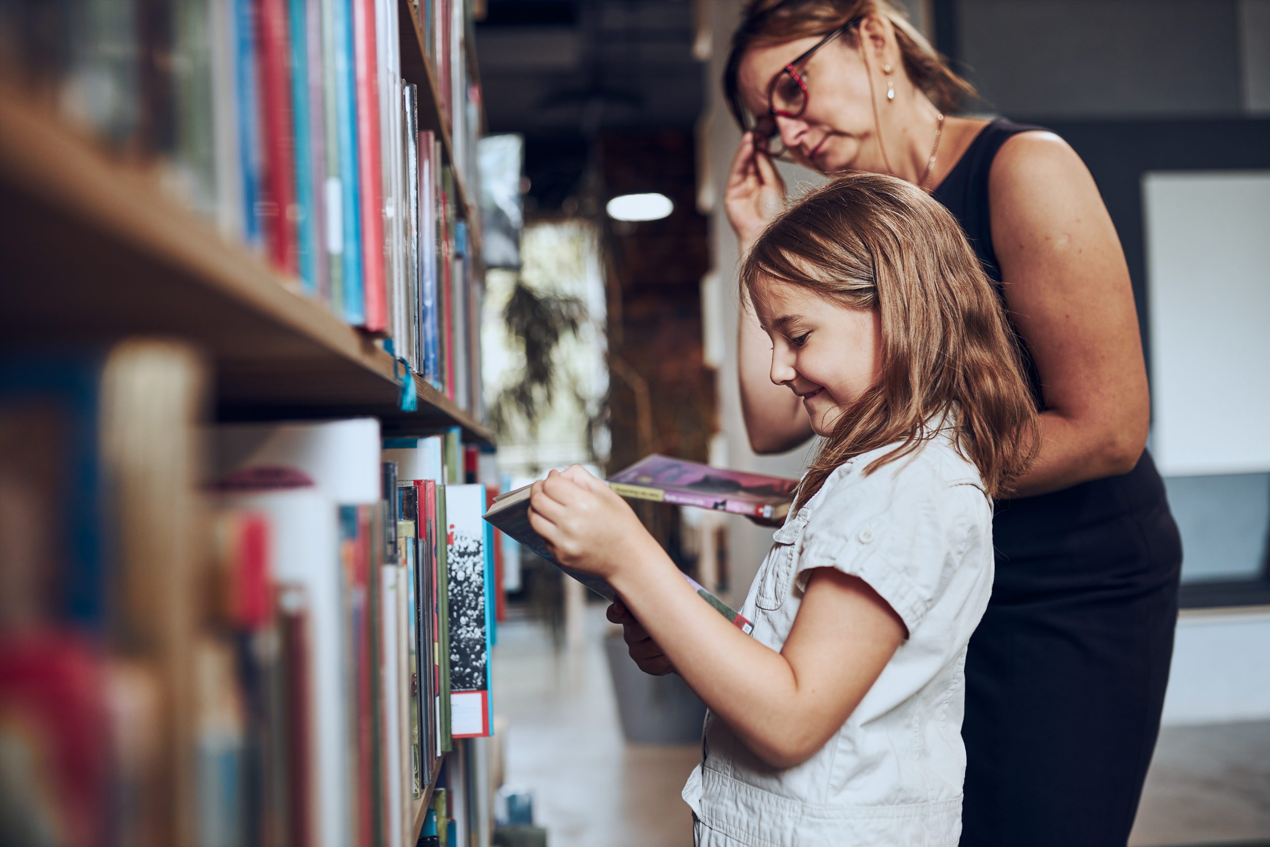 teacher helping to choose book her schoolgirl in school library. smart girl selecting literature for reading. books on shelves in bookstore. learning from books. school education. benefits of everyday reading. child curiosity